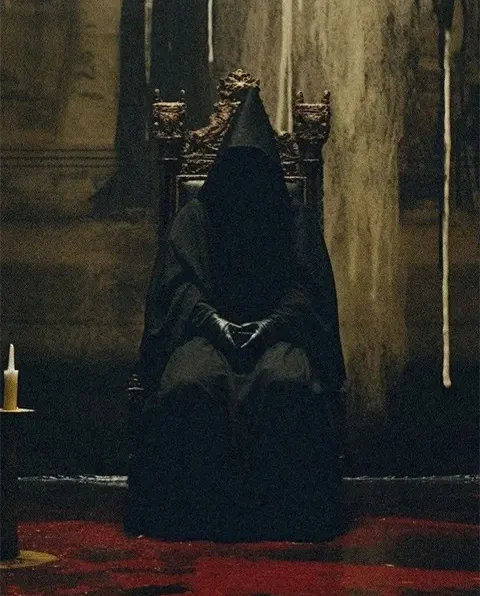 Dark figure wearing black cap seated on throne